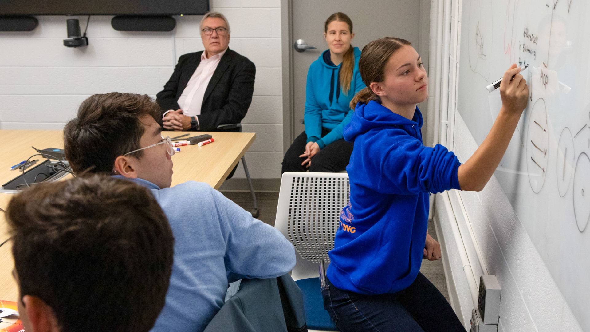 Instructors and students watch as one student writes an equation on a whiteboard.